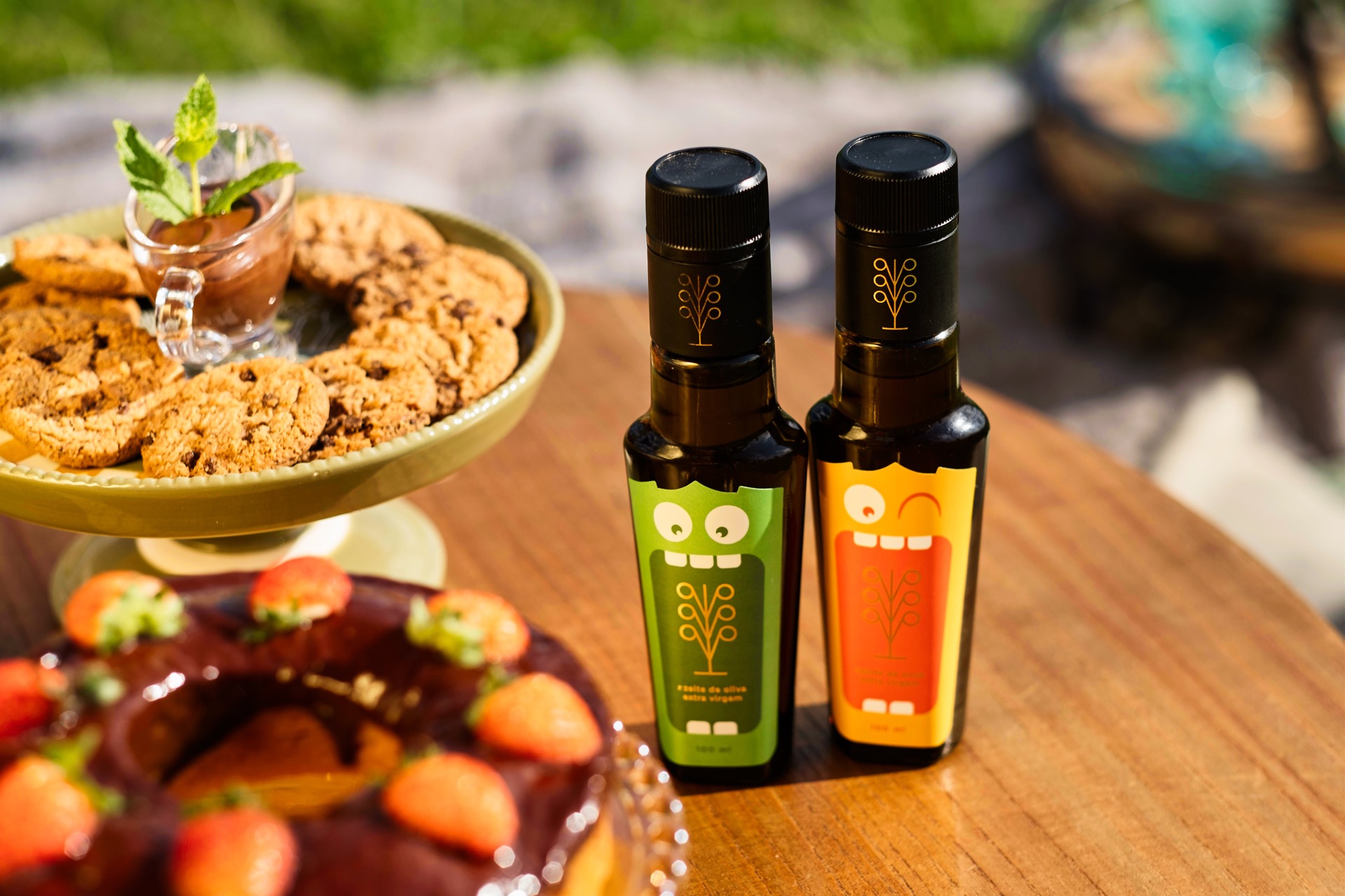 Duas garrafinhas Azito's com os rótulos monstrinho verde e laranja sobre mesa de madeira, ao lado de um prato de biscoitos e de um bolo de chocolate com morangos.