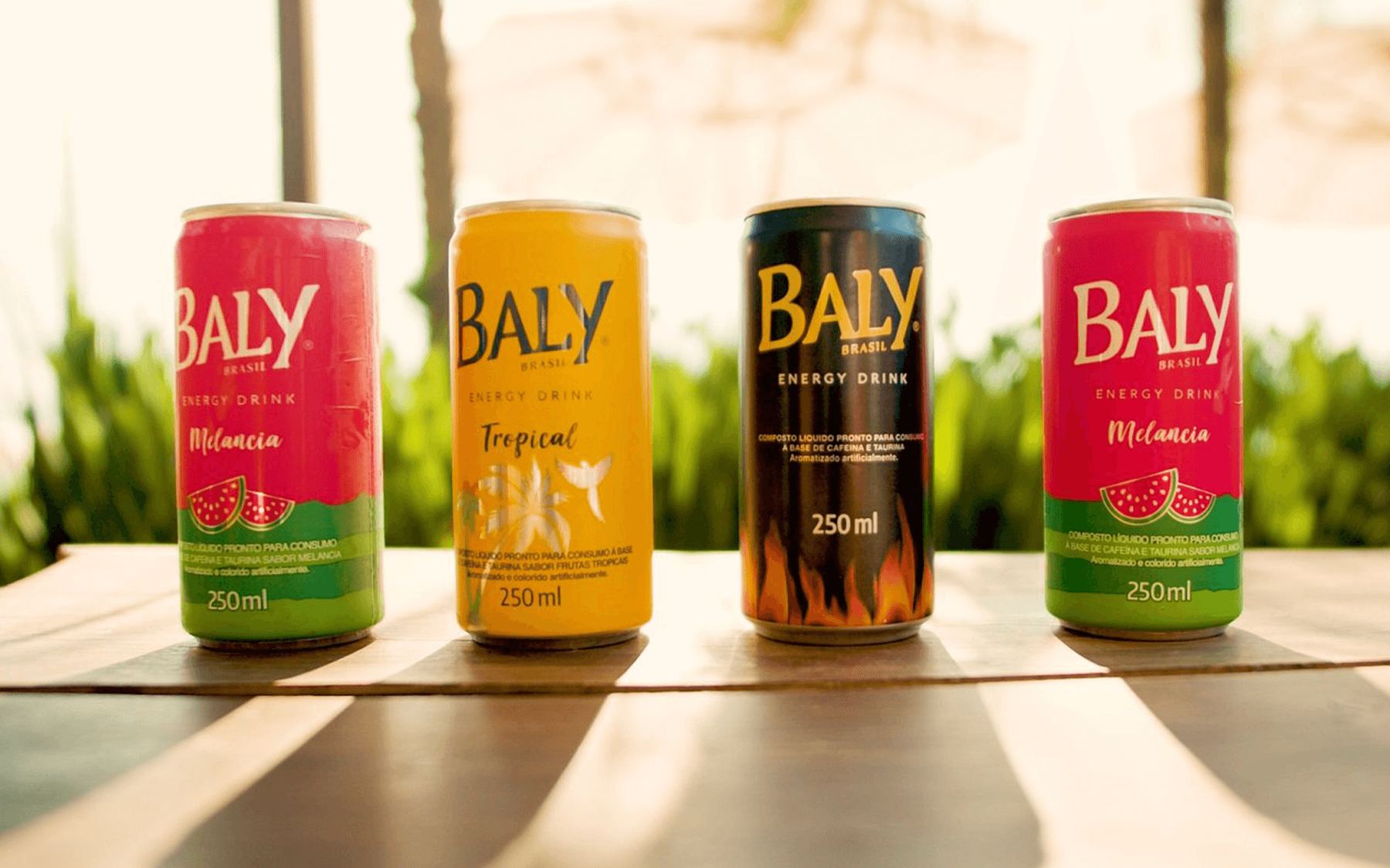 Quatro latas de energético Baly de sabores diferentes enfileiradas sobre uma mesa de madeira.