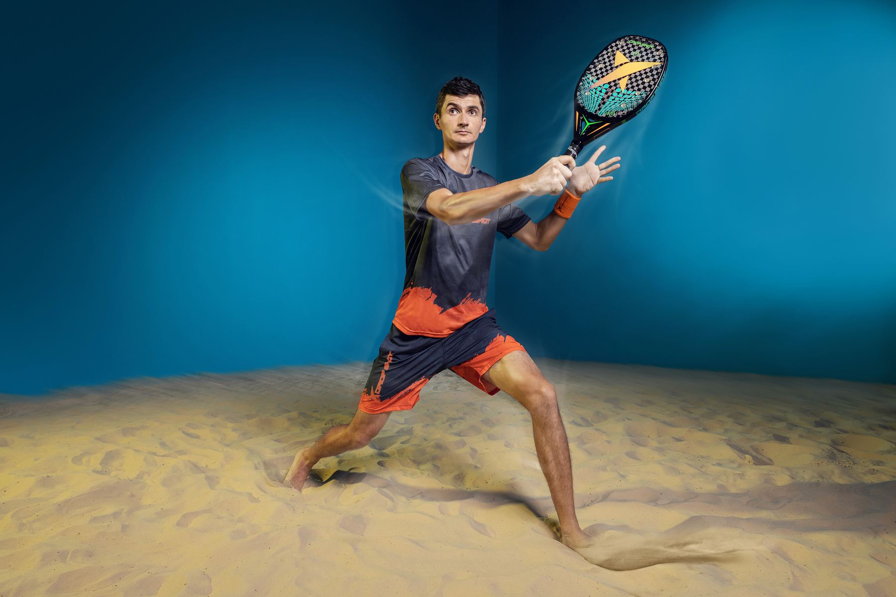 Atleta de beach tennis rebatendo sobre a areia, com rastro de movimento e fundo azul.