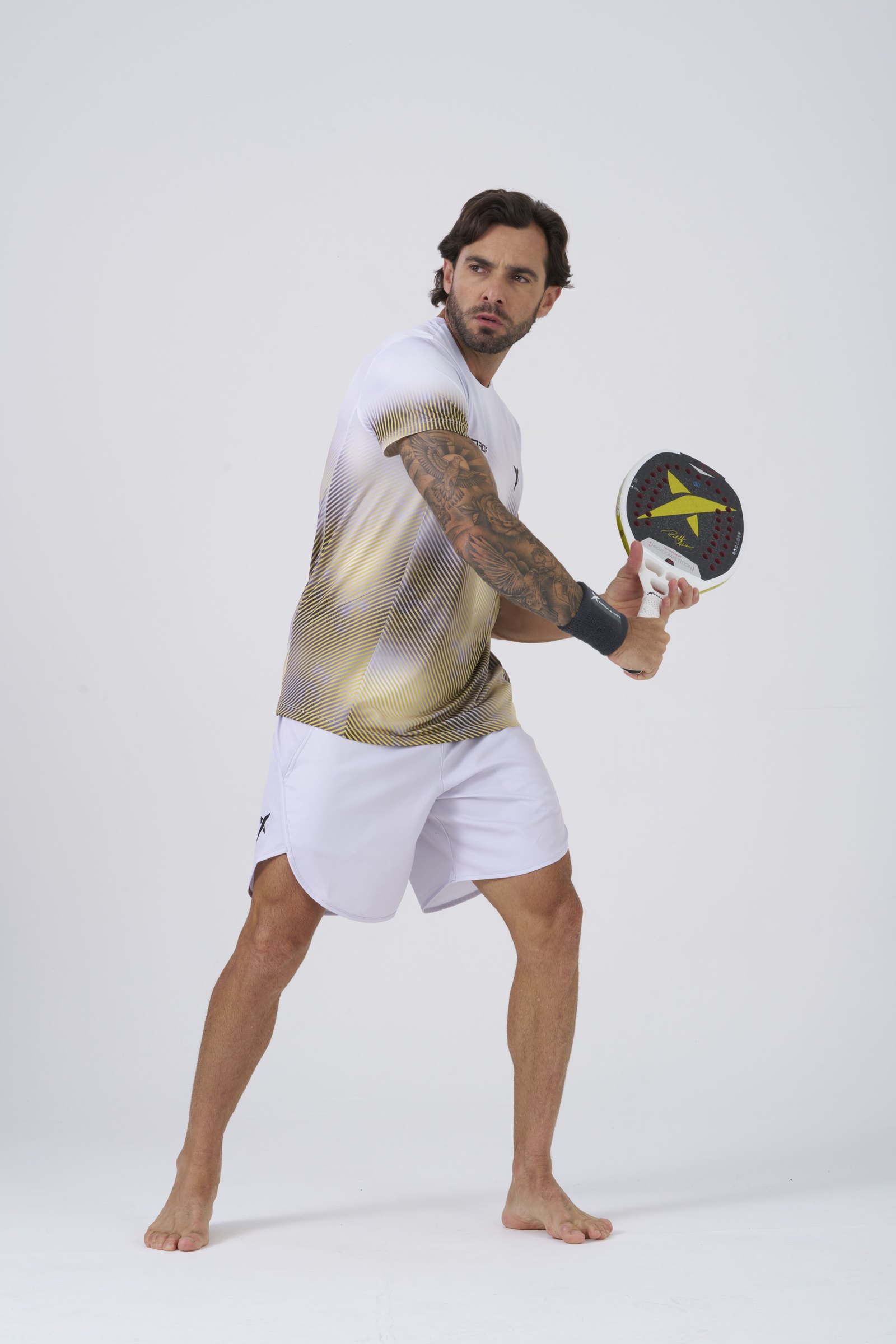 Modelo de camiseta e short brancos da Drop Shot em movimento com a raquete, em estúdio claro.