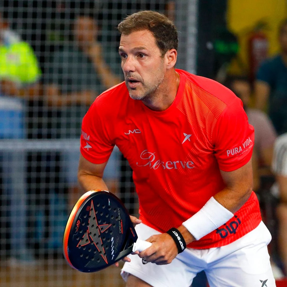 Jogador de padel de camiseta vermelha, em posição de espera na quadra, com a raquete Drop Shot nas mãos.