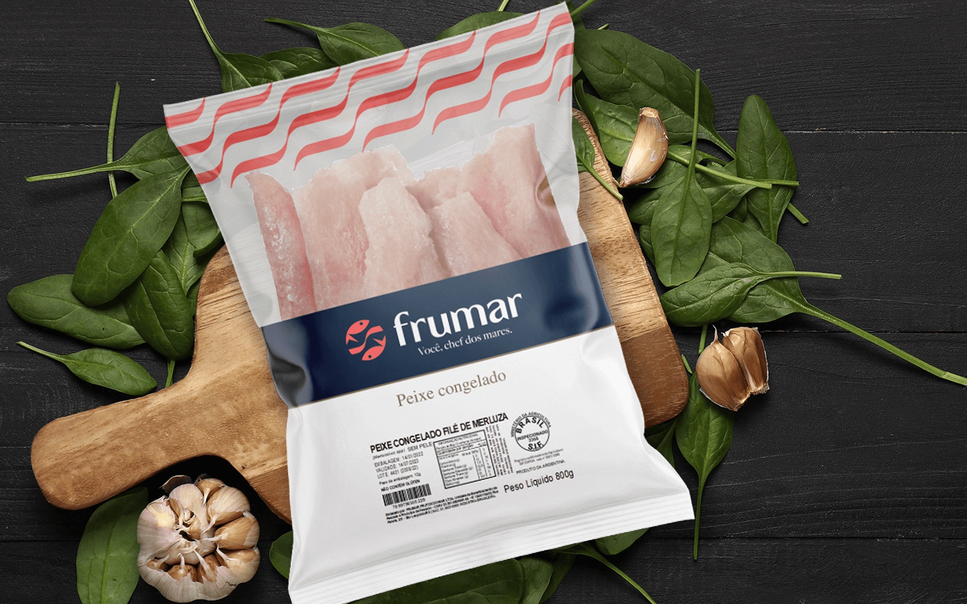Embalagem Frumar de peixe congelado filé de merluza sobre tábua de madeira, cercada por folhas de espinafre e dentes de alho, em fundo de madeira preta.