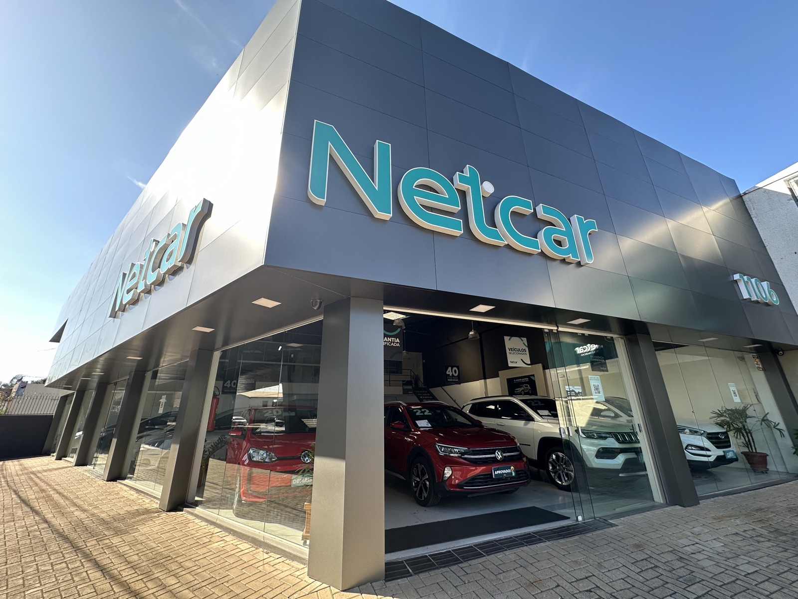 Fachada da Netcar: letreiro turquesa no prédio cinza e seminovos atrás da vitrine de vidro.