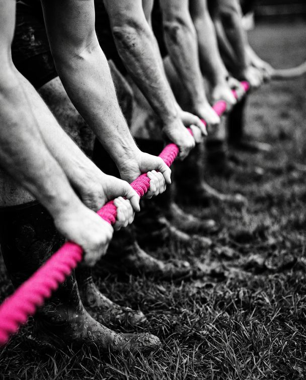 Fila de mãos puxando a mesma corda rosa num cabo de guerra, foto em preto e branco.