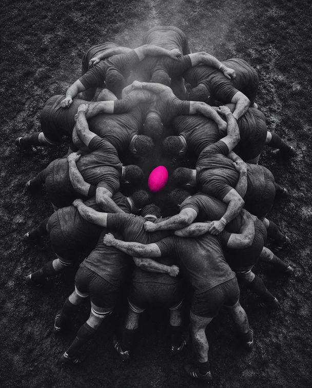 Scrum de rugby visto de cima: jogadores abraçados num bloco só, com a bola rosa no centro.