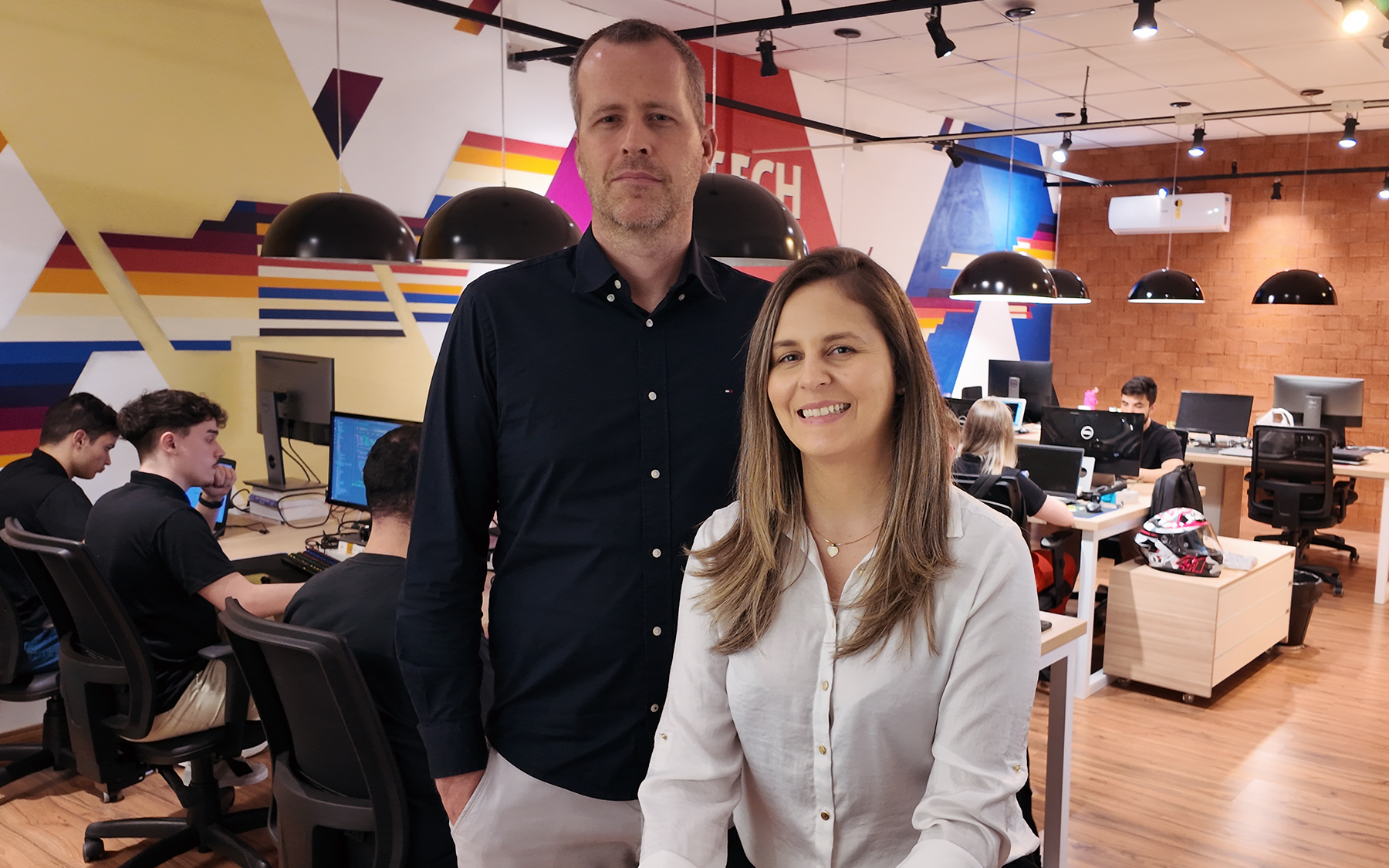 Um homem e uma mulher posam no escritório da 3035TECH, com a equipe trabalhando nos computadores ao fundo e um mural geométrico colorido na parede