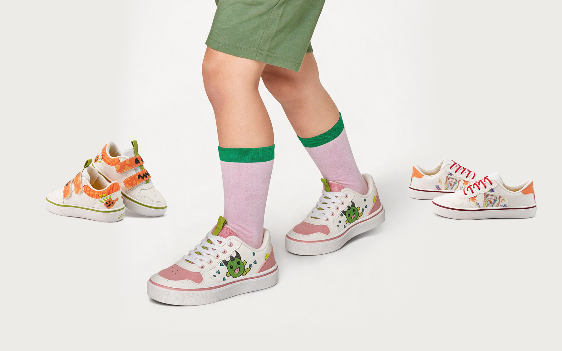 Criança de meias cor-de-rosa calçando tênis brancos da Linha Monstrinhos, com um monstrinho verde estampado, ao lado de outros pares da coleção.